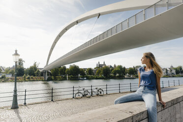 Netherlands, Maastricht, young woman sitting on a wall at the riverside - GUSF01527