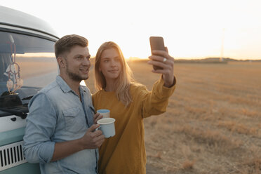 Young couple taking a selfie at camper van in rural landscape at sunset - GUSF01539