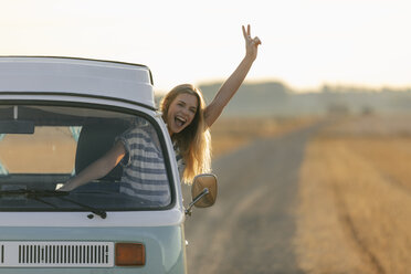 Excited young woman making victory hand sign out of camper van window in rural landscape - GUSF01548
