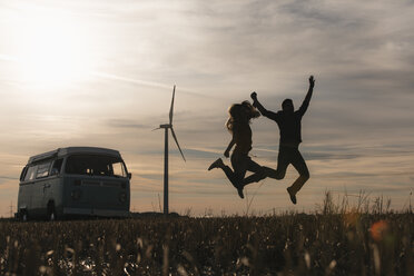 Exuberant couple jumping at camper van in rural landscape at dusk - GUSF01556