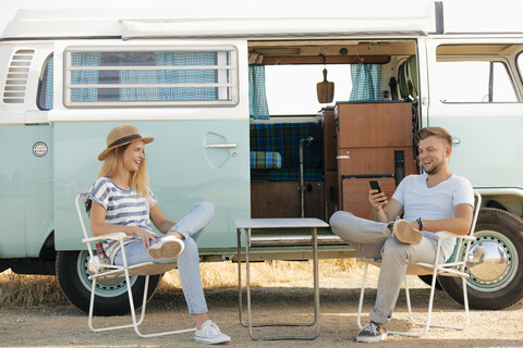 Glückliches junges Paar auf Campingstühlen im Wohnmobil sitzend mit Mobiltelefon, lizenzfreies Stockfoto