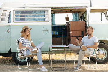 Glückliches junges Paar auf Campingstühlen im Wohnmobil sitzend mit Mobiltelefon - GUSF01577