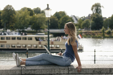 Netherlands, Maastricht, young woman sitting on a wall at the riverside with laptop - GUSF01612