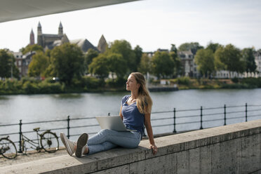 Niederlande, Maastricht, junge Frau sitzt mit Laptop auf einer Mauer am Flussufer - GUSF01613