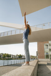 Netherlands, Maastricht, young woman standing on a wall at the riverside with raised arms - GUSF01622