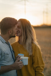 Happy young couple holding mugs kissing in rural landscape - GUSF01631