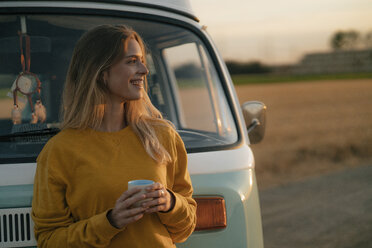 Happy young woman holding mug at camper van in rural landscape at sunset - GUSF01633