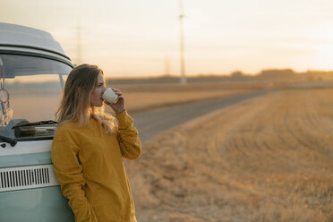 Young woman drinking from mug at camper van in rural landscape at sunset - GUSF01634