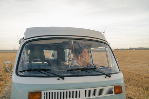 Lächelnde junge Frau am Steuer eines Wohnmobils in ländlicher Landschaft, lizenzfreies Stockfoto