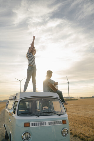 Glückliches Paar mit Gitarre auf dem Dach eines Wohnmobils in ländlicher Landschaft, lizenzfreies Stockfoto