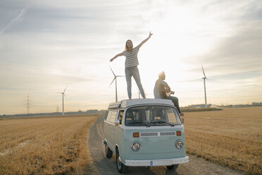 Happy couple with guitar on roof of a camper van in rural landscape - GUSF01645