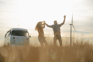 Exuberant couple at camper van in rural landscape with wind turbine in background - GUSF01647