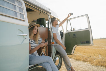 Young couple with cell phones relaxing at camper van in rural landscape - GUSF01658