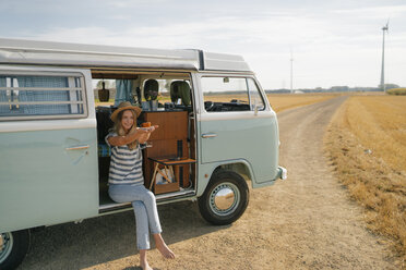 Smiling young woman holding cup of coffee in camper van in rural landscape - GUSF01660