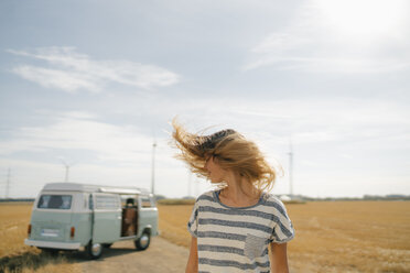 Blonde junge Frau am Wohnmobil in ländlicher Landschaft schüttelt ihr Haar - GUSF01664