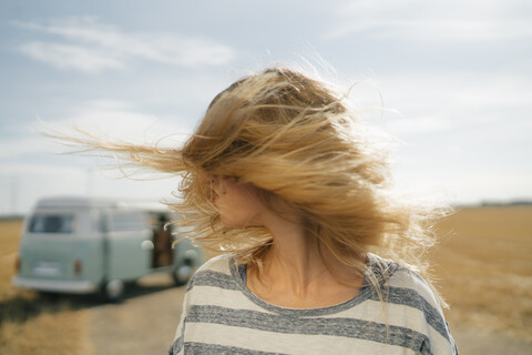 Blonde junge Frau am Wohnmobil in ländlicher Landschaft schüttelt ihr Haar, lizenzfreies Stockfoto