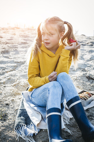 Porträt eines Mädchens mit Zöpfen, das im Herbst am Strand sitzt und einen Apfel isst, lizenzfreies Stockfoto