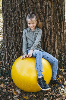 Porträt eines lächelnden Mädchens, das auf einem gelben Gymnastikball sitzt, der an einem Baumstamm lehnt - HMEF00091