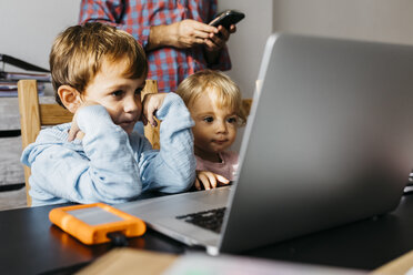 Bruder und Schwester sehen sich ein Video auf einem Laptop an, der Vater benutzt ein Smartphone im Hintergrund - JRFF01986