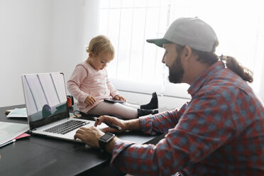 Vater, der von zu Hause aus arbeitet und einen Laptop benutzt, während seine Tochter auf dem Schreibtisch sitzt und spielt - JRFF02008