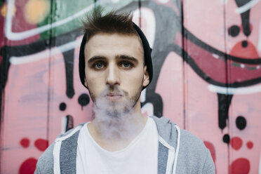 Young man standing in front of graffiti, smoking electronic cigarette - JRFF02030