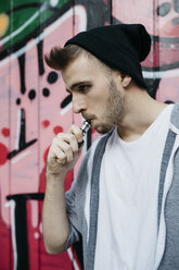 Junger Mann steht vor einem Graffiti und raucht eine elektronische Zigarette - JRFF02031
