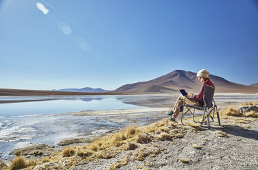 Bolivien, Laguna Colorada, Frau sitzt auf Campingstuhl am Seeufer und benutzt Tablet - SSCF00029