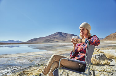 Bolivien, Laguna Colorada, Frau sitzt auf Campingstuhl am Seeufer und trinkt aus Becher - SSCF00030