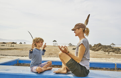 Chile, Arica, glückliche Mutter in Yogastellung mit Sohn an der Wand sitzend am Strand, lizenzfreies Stockfoto