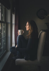 Nachdenkliche Frau mit Kaffeetasse, die durch das Fenster schaut, während sie zu Hause sitzt - CAVF55884