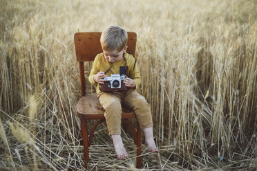 Full length of boy with vintage camera sitting on chair amidst wheat field - CAVF55910