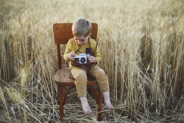 Junge mit Vintage-Kamera in voller Länge auf einem Stuhl inmitten eines Weizenfeldes sitzend - CAVF55910