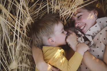 High angle view of playful son biting mother's fingers while lying on wheat field - CAVF55922