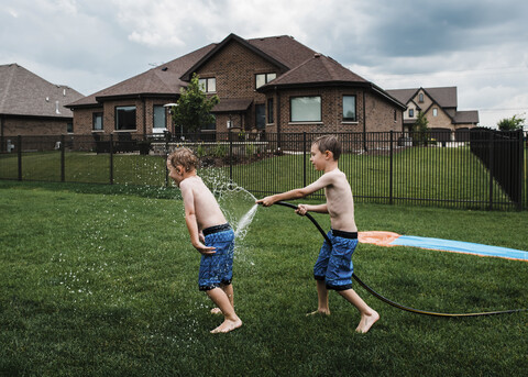 Side view of shirtless boy spraying water on brother with garden hose at park stock photo