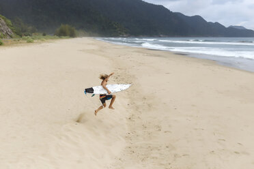 Hohe Winkelansicht des hemdlosen Mannes mit Surfbrett, der am Strand springt - CAVF55934