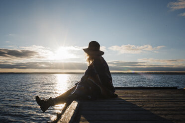 Seitenansicht einer in eine Decke eingewickelten Frau, die während des Sonnenuntergangs auf einem Pier am Lake Simcoe sitzt - CAVF55977