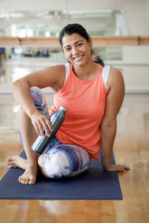 Porträt einer lächelnden Frau, die eine Flasche hält, während sie im Fitnessstudio sitzt - CAVF55992