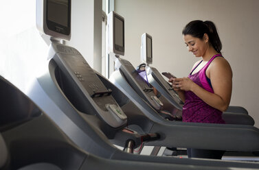 Lächelnde Frau, die ein Smartphone benutzt, während sie auf dem Laufband im Fitnessstudio steht - CAVF56008