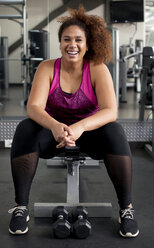 Porträt einer fröhlichen Frau, die auf einer Bank im Fitnessstudio sitzt - CAVF56009