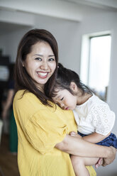 Porträt einer japanischen Frau, die in einem Wohnzimmer steht und ein junges Mädchen trägt, das in die Kamera lächelt. - MINF09586