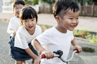 Porträt von zwei japanischen Mädchen und einem Jungen, die auf der Straße mit einem Fahrrad spielen und in die Kamera lächeln. - MINF09594