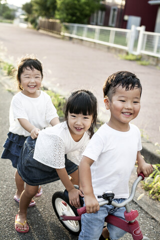 Porträt von zwei japanischen Mädchen und einem Jungen, die auf der Straße mit einem Fahrrad spielen und in die Kamera lächeln., lizenzfreies Stockfoto