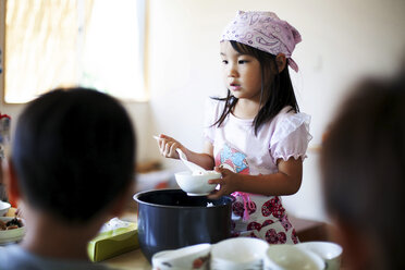 Ein Mädchen mit Kopftuch steht an einem Tisch in einer japanischen Vorschule und serviert den Kindern das Mittagessen. - MINF09625
