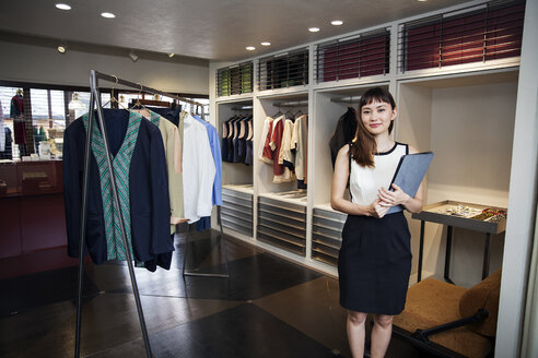 Lächelnde japanische Verkäuferin in einem Bekleidungsgeschäft, die einen Ordner hält und in die Kamera lächelt. - MINF09668