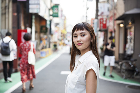 Lächelnde Japanerin mit langen braunen Haaren und weißer kurzärmeliger Bluse steht auf einer Straße. - MINF09671