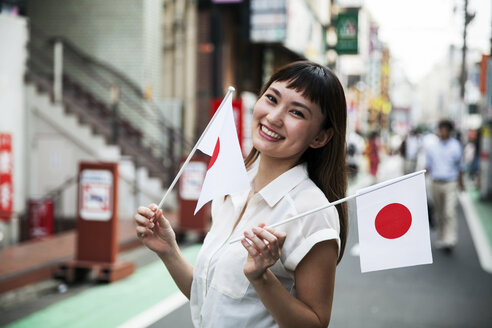 Lächelnde Japanerin mit langen braunen Haaren und weißer kurzärmeliger Bluse steht auf einer Straße und hält eine kleine japanische Flagge in der Hand. - MINF09672