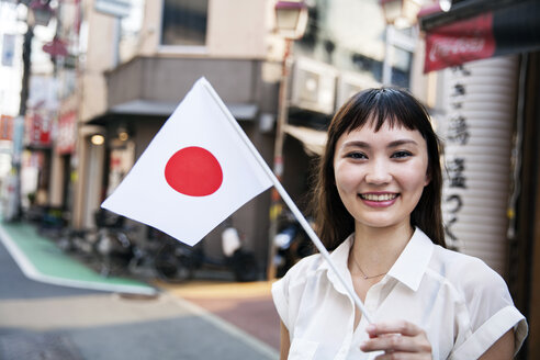 Lächelnde Japanerin mit langen braunen Haaren und weißer kurzärmeliger Bluse steht auf einer Straße und hält eine kleine japanische Flagge in der Hand. - MINF09673