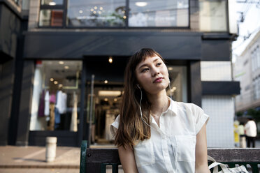 Japanese woman with long brown hair wearing white short-sleeved blouse sitting on a bench, wearing earphones, listening to music. - MINF09674