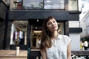Japanische Frau mit langen braunen Haaren und weißer kurzärmeliger Bluse sitzt auf einer Bank, trägt Kopfhörer und hört Musik. - MINF09674