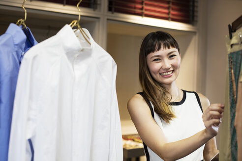Lächelnde japanische Verkäuferin, die in einem Bekleidungsgeschäft steht und ein Hemd betrachtet. - MINF09677